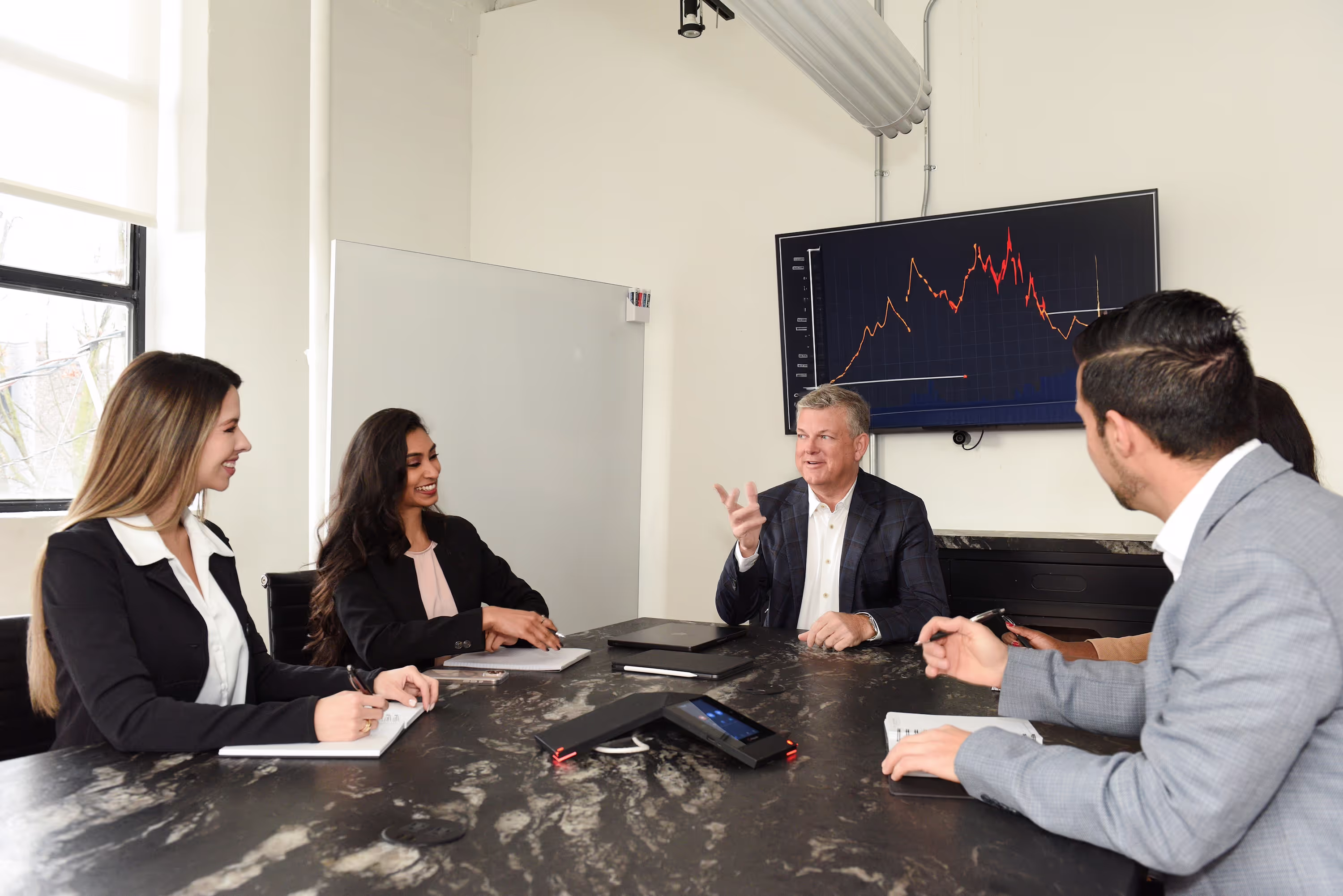 Group of people sitting around an office table, discussing ideas. A large TV screen in the background displays a graph with data analysis.
